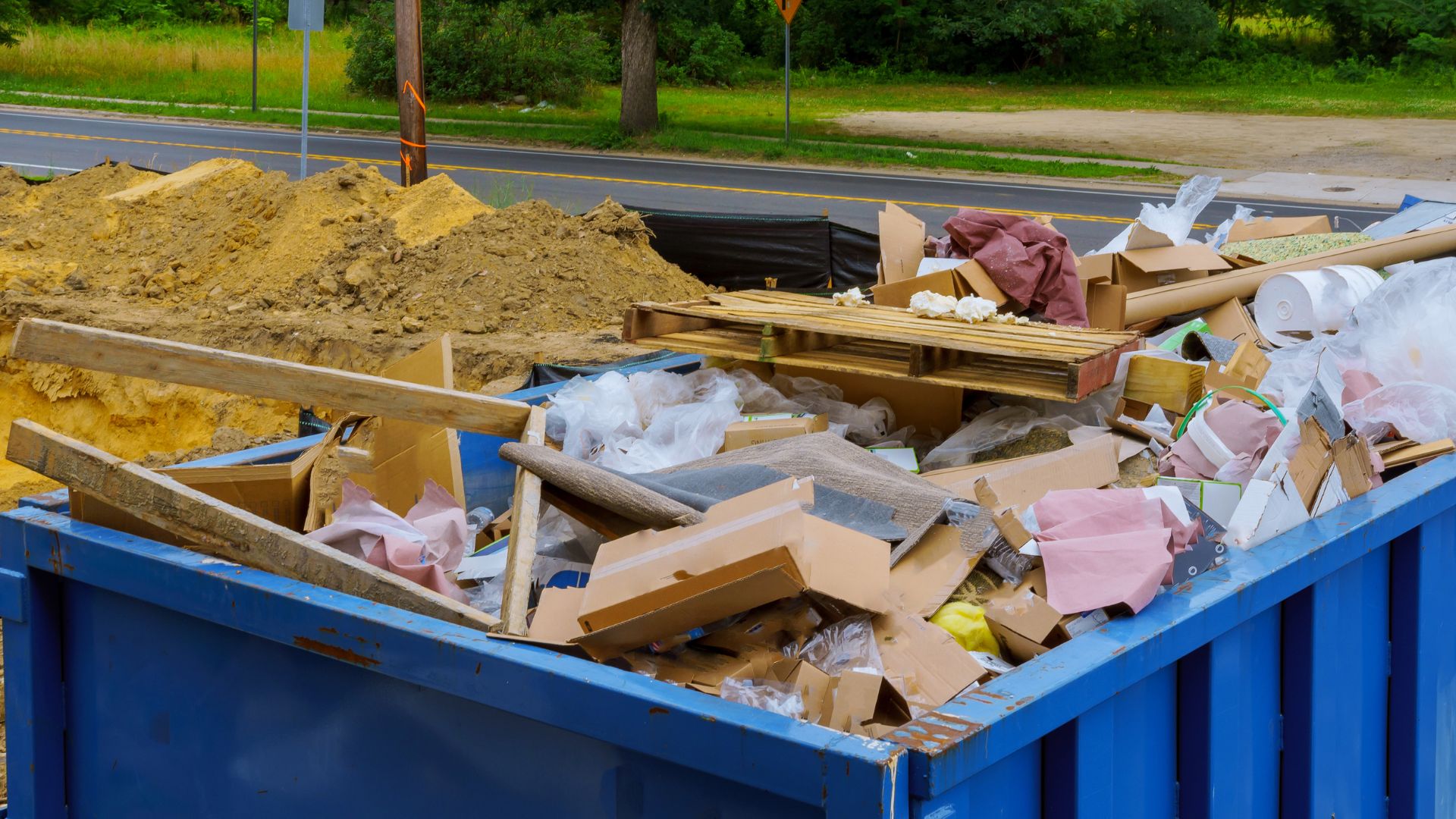 Blue dumpster filled with construction debris, wood, and cardboard near roadside